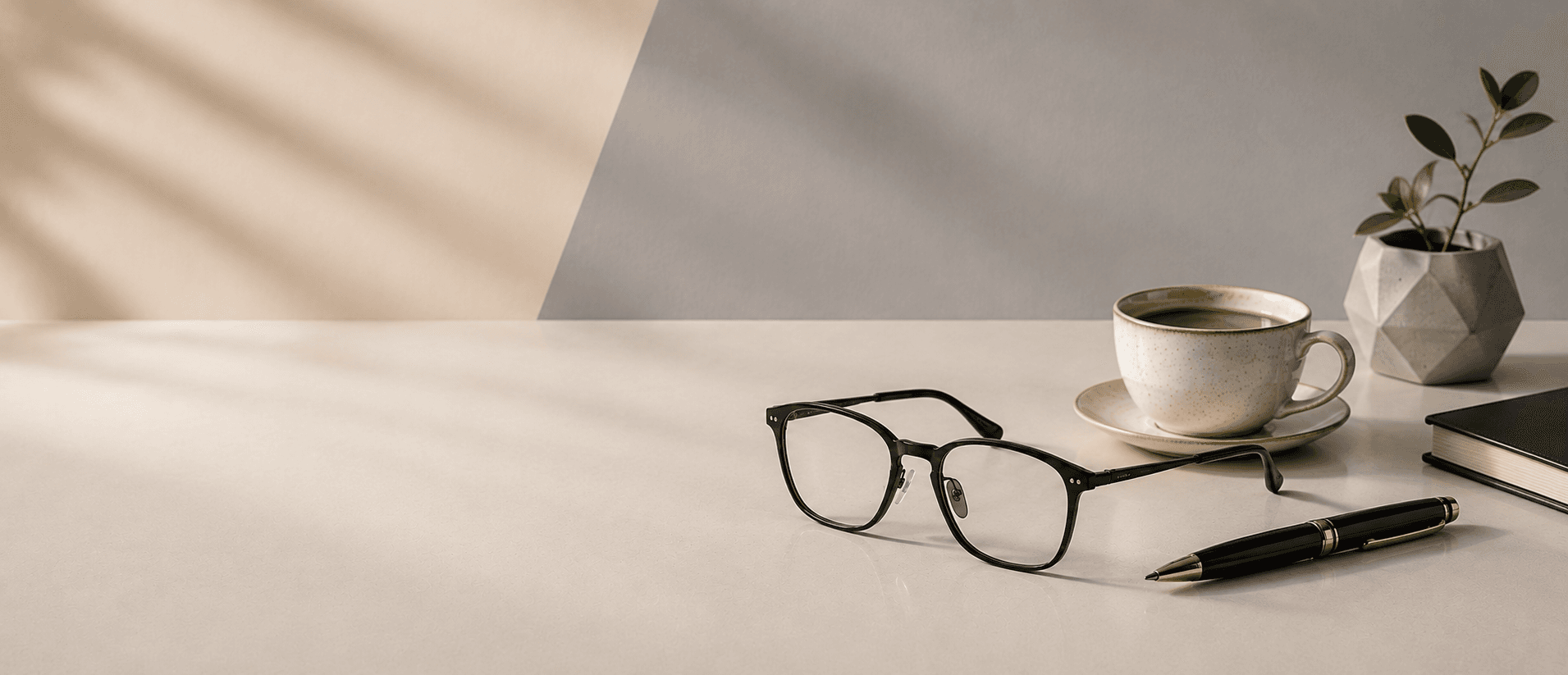 Black eyeglasses on a desk beside a cup of coffee, a pen and a small plant